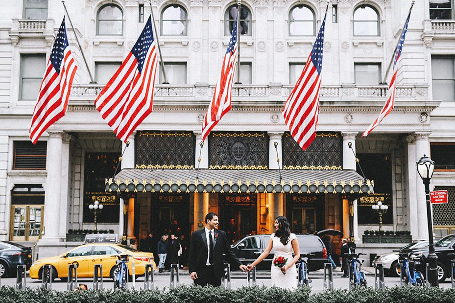 A couple stands in front of the Plaza Hotel in New York City. The woman is wearing a wedding dress and the man is wearing a suit. They are holding hands and smiling. The United States flag is flying in the background. This image is for a blog post about how to get married in New York City. The couple used our guide to help them get their marriage license.